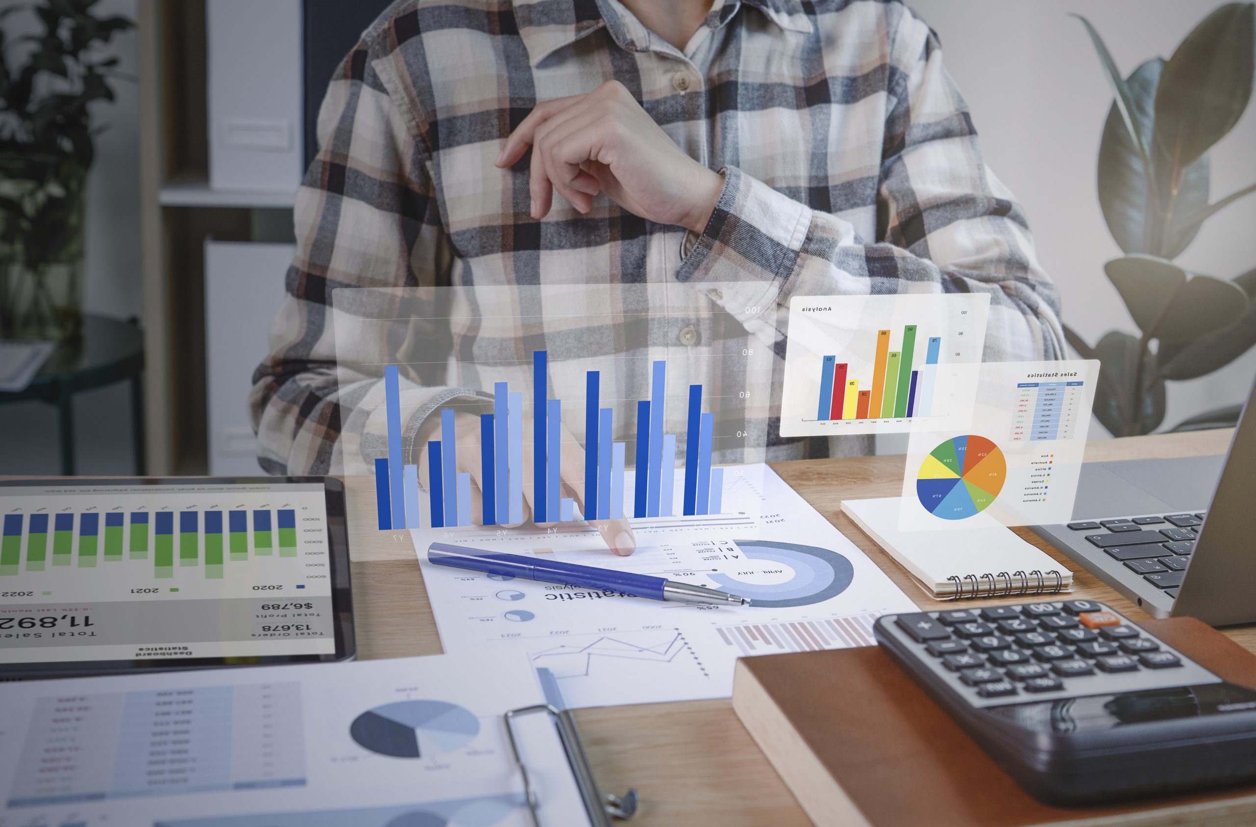 Profissional analisando dados financeiros em um ambiente de trabalho moderno. Sobre a mesa, há gráficos impressos, uma calculadora, um laptop e um tablet exibindo estatísticas. Elementos gráficos digitais flutuantes representam informações visuais, como gráficos de barras e pizza, sugerindo o uso de tecnologia avançada para análise de dados e tomada de decisões estratégicas.