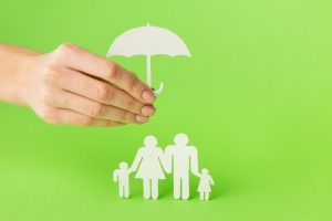 Uma mão segura um guarda-chuva de papel branco sobre uma família representada por recortes de papel, composta por dois adultos e duas crianças, simbolizando proteção e segurança. O fundo verde vibrante destaca os elementos brancos, reforçando a ideia de seguro de vida, estabilidade financeira e bem-estar familiar.