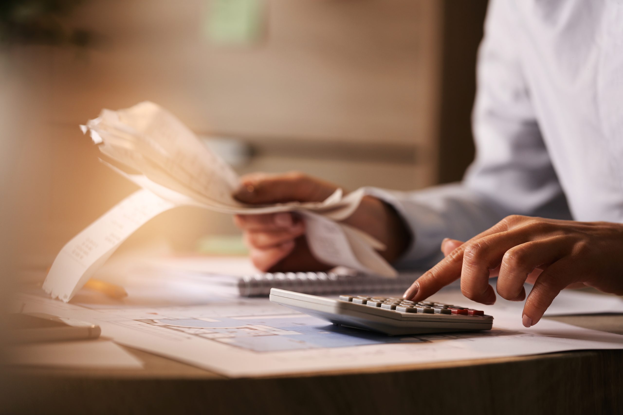 Close de uma pessoa analisando recibos e digitando em uma calculadora. A cena sugere a revisão de contas, impostos ou planejamento financeiro. O ambiente tem iluminação suave e documentos espalhados sobre a mesa, transmitindo um clima de concentração e organização.