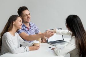 Um jovem casal sorridente está em uma reunião com uma consultora financeira. O homem aperta a mão da profissional, demonstrando satisfação e confiança, enquanto a mulher, segurando um copo de café, observa com um sorriso. Sobre a mesa, há um laptop, uma caneta e documentos, indicando um ambiente de negociação ou assinatura de contrato.