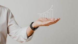 Uma mão estendida, vestida com uma camisa social branca e um relógio preto, exibe um gráfico digital crescente projetado no ar. O gráfico representa um crescimento contínuo, simbolizando sucesso financeiro, progresso empresarial e estratégias eficazes para aumentar os resultados. A imagem transmite inovação, planejamento estratégico e desenvolvimento no ambiente corporativo.