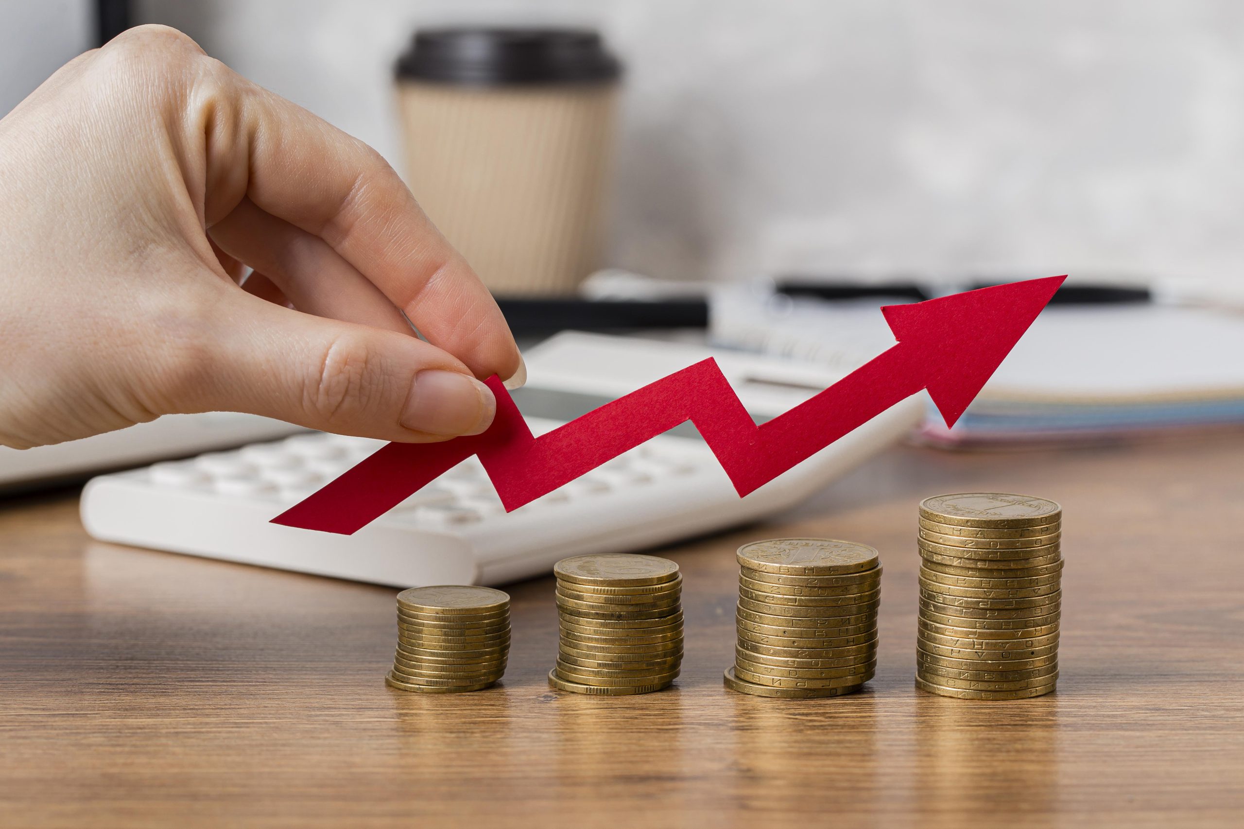 Uma mão segura uma seta vermelha em forma de gráfico ascendente sobre pilhas de moedas empilhadas em ordem crescente. Ao fundo, há uma calculadora, um caderno e uma xícara de café, sugerindo um ambiente de planejamento financeiro. A imagem simboliza crescimento econômico, aumento de lucros e estratégias de investimento para o sucesso financeiro.