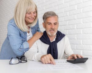 Casal de idosos analisa juntos um documento financeiro sobre uma mesa branca; o homem, sentado, aponta para o papel enquanto segura uma calculadora, e a mulher, em pé ao seu lado, sorri com a mão sobre o ombro dele, transmitindo cumplicidade e planejamento conjunto.