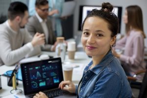 Uma mulher sorridente, vestindo uma jaqueta jeans, está sentada em um ambiente corporativo com um laptop aberto exibindo gráficos e análises de dados. Ao fundo, um grupo de colegas participa de uma reunião, discutindo informações em um escritório moderno. A imagem transmite um ambiente de trabalho colaborativo e tecnológico, destacando a importância da análise de dados e do trabalho em equipe no mundo corporativo.