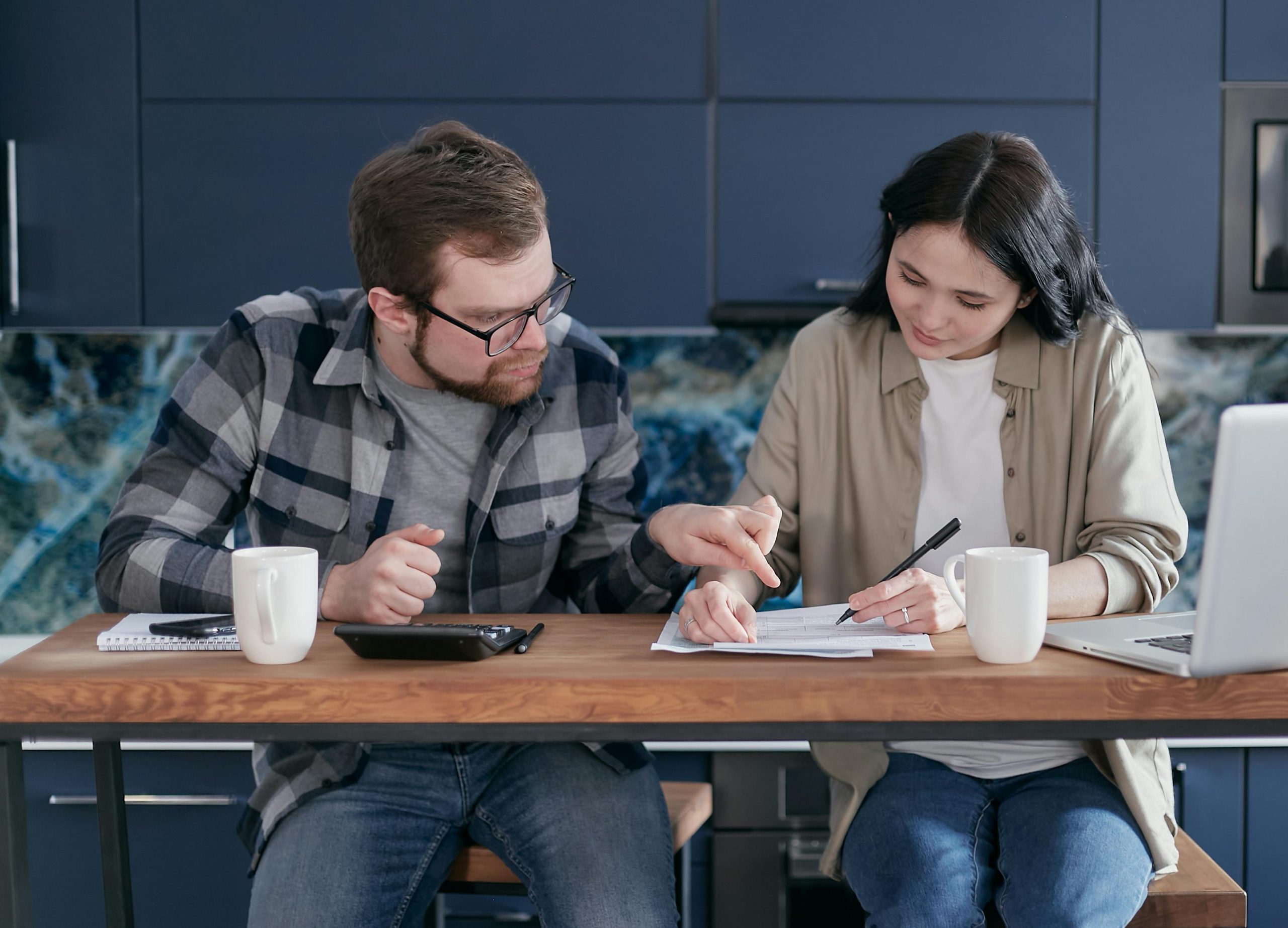 Casal sentado à mesa da cozinha analisa documentos financeiros com concentração; o homem aponta para um trecho do papel enquanto a mulher faz anotações com uma caneta, acompanhados por uma calculadora, dois copos e um notebook, em um ambiente moderno e organizado.