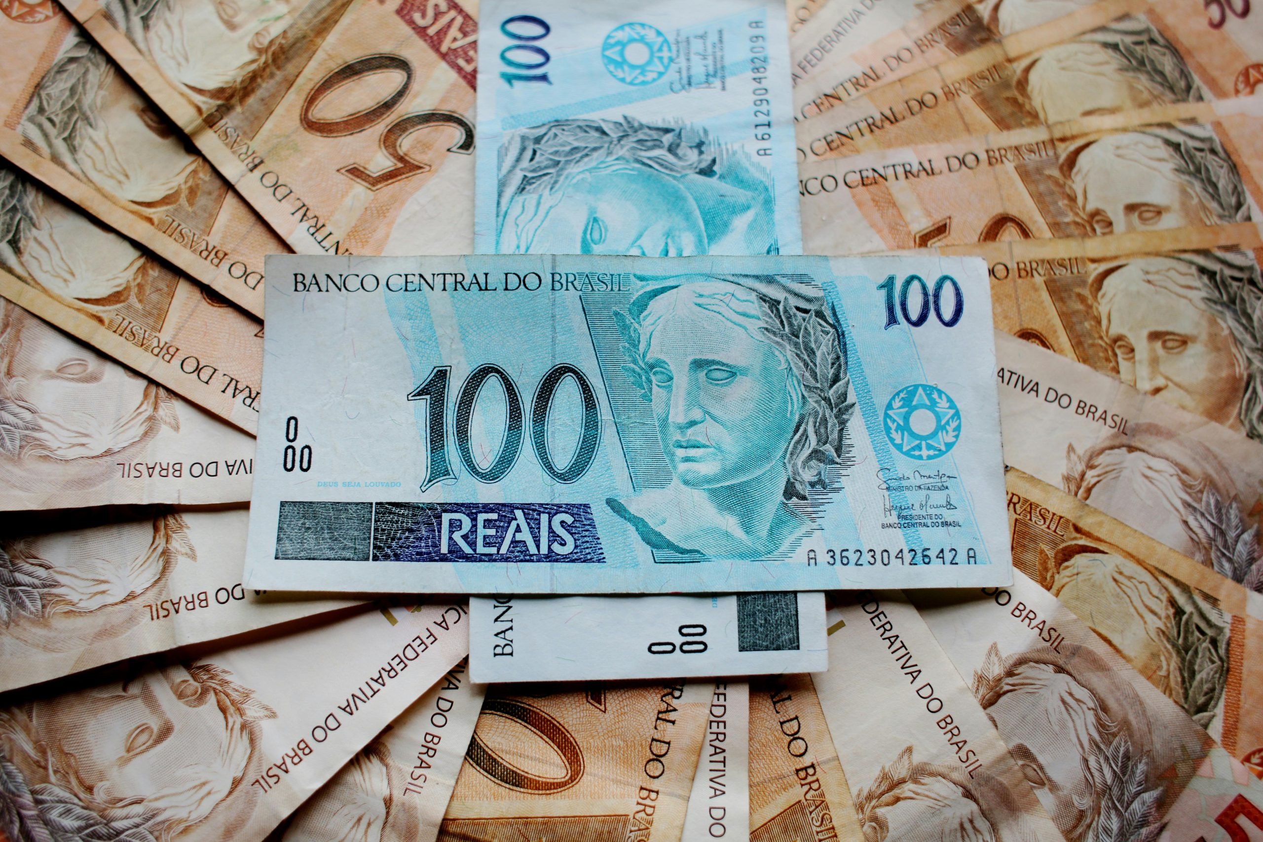 Notas de real brasileiro dispostas em formato de leque, com destaque para uma cédula de R$ 100 posicionada no centro, sobre outras notas de R$ 50 e R$ 100 parcialmente visíveis, representando riqueza, economia ou finanças no Brasil.
