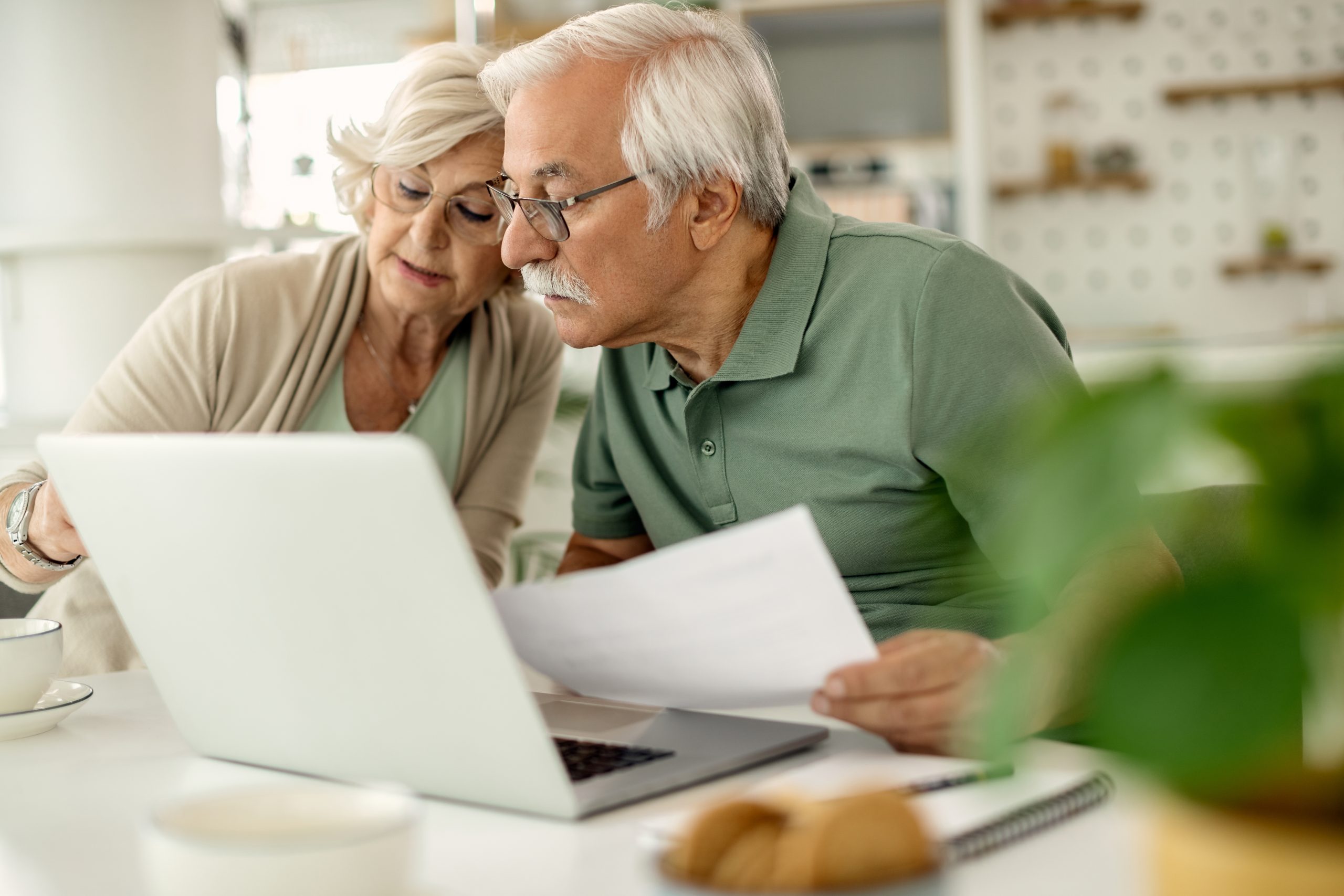 Casal de idosos sentado à mesa em casa, concentrado na tela de um notebook enquanto analisa documentos financeiros. Ambos demonstram atenção e cuidado ao revisar suas contas e planejamentos. A cena ilustra a importância da finanças pessoais para aposentados, reforçando a necessidade de organização, planejamento e tomada de decisões conscientes para garantir estabilidade e qualidade de vida na terceira idade.