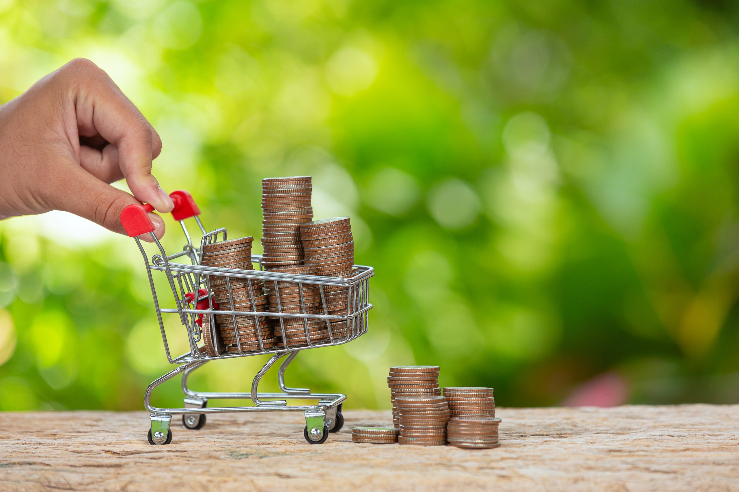 Mão empurrando um carrinho de compras em miniatura cheio de moedas empilhadas, com mais moedas empilhadas ao lado sobre uma superfície de madeira. Ao fundo, há uma paisagem desfocada de natureza em tons de verde. A imagem representa os impactos da inflação no poder de compra, simbolizando como o valor do dinheiro é afetado e como os consumidores precisam ajustar seus gastos frente à desvalorização monetária.