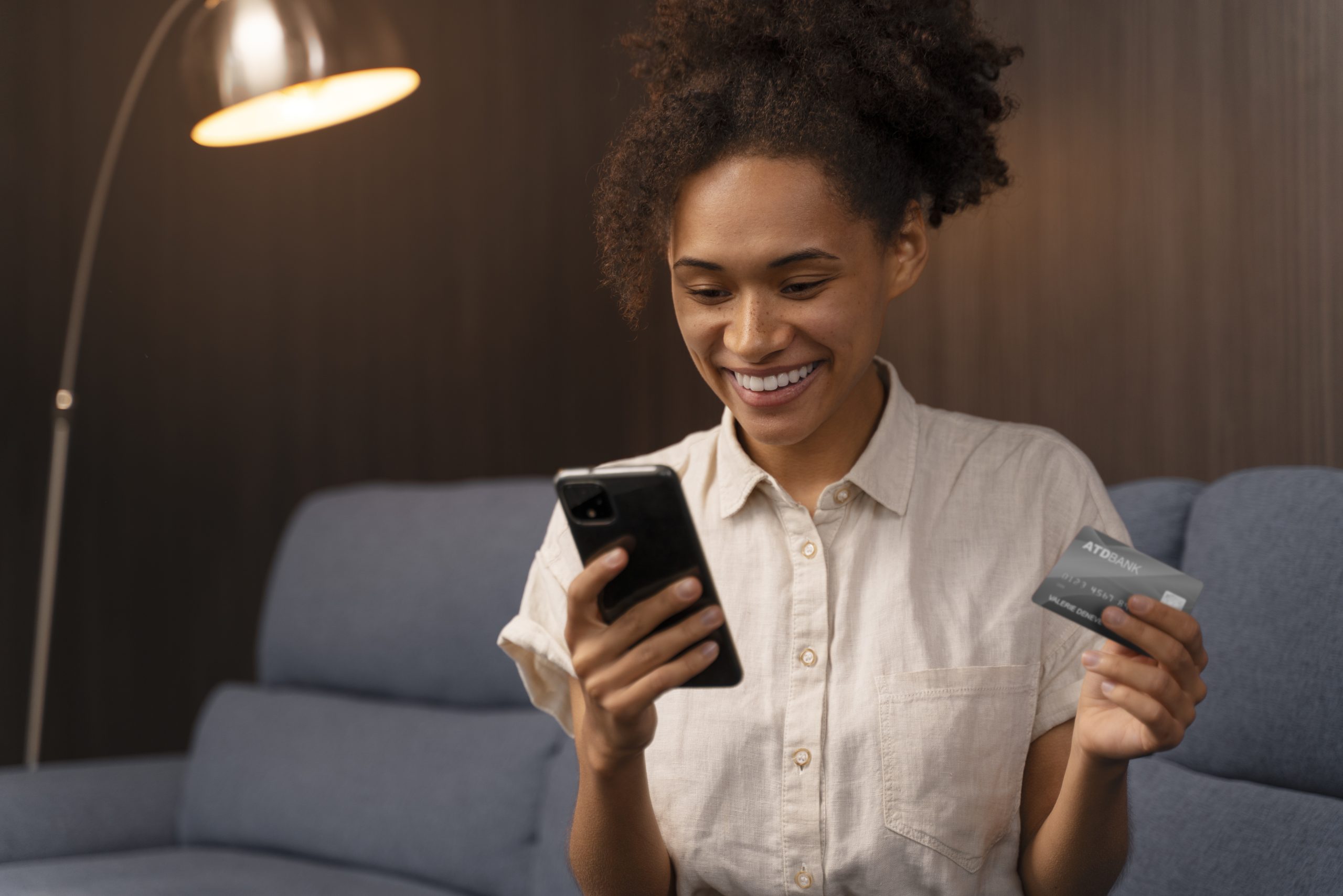 Mulher sorridente sentada em um sofá utilizando um smartphone enquanto segura um cartão de crédito na outra mão. Ao fundo, há uma luminária acesa e uma parede de madeira escura, criando um ambiente acolhedor. A cena representa a praticidade e conveniência do crédito digital, evidenciando como as soluções financeiras modernas facilitam o acesso a compras e serviços de forma rápida e segura.