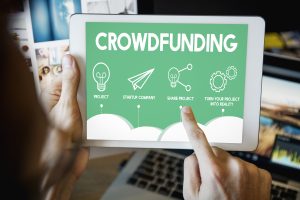 Pessoa segurando um tablet que exibe uma tela verde com o título "Crowdfunding" e ícones representando as etapas do financiamento coletivo: criação de projeto, abertura de startup, compartilhamento do projeto e transformação da ideia em realidade, enquanto um dedo aponta para a etapa "Share Project".