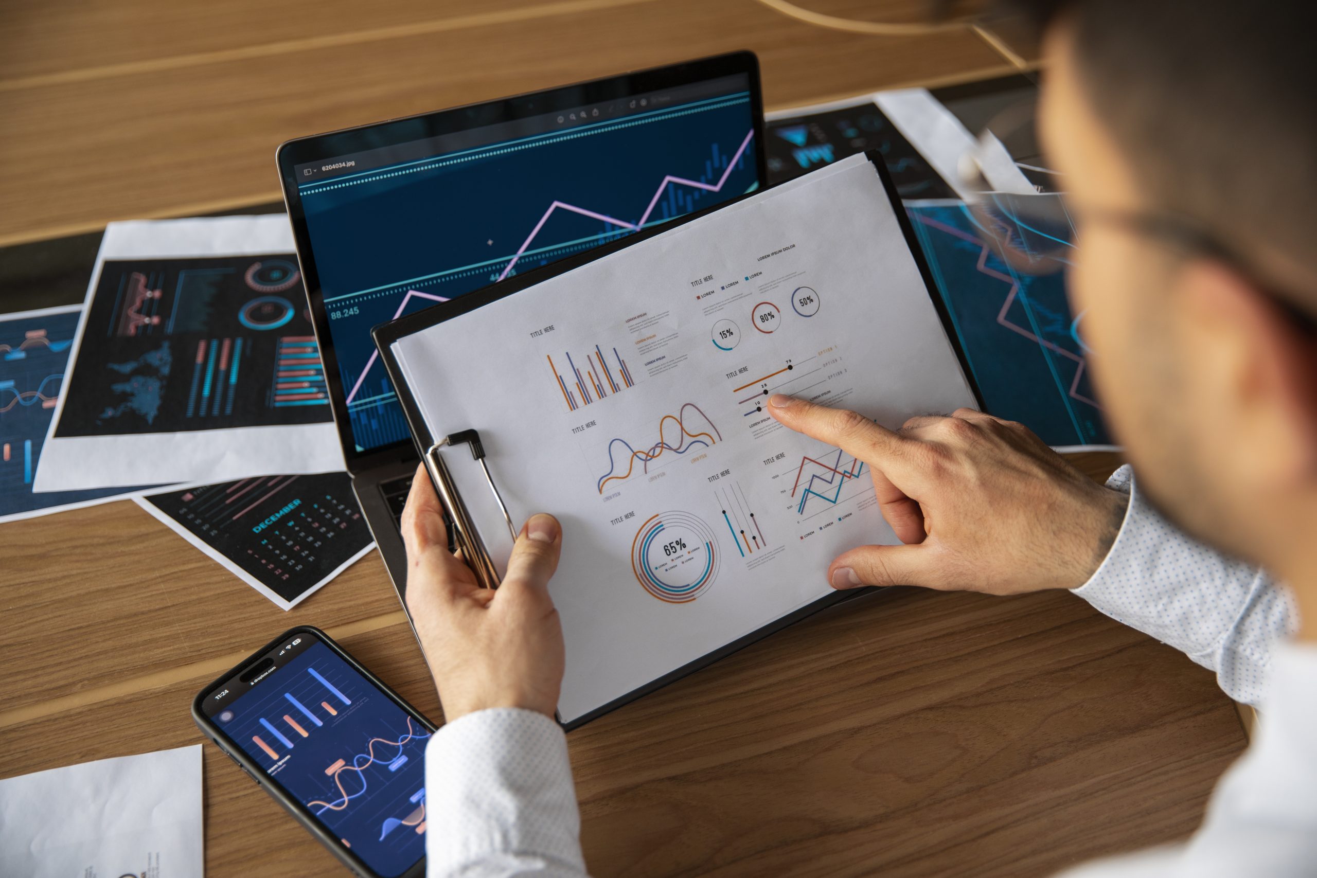 Profissional analisando gráficos e relatórios financeiros em papel, enquanto compara com dados exibidos na tela de um notebook e de um smartphone, ambos sobre uma mesa de madeira. O ambiente está repleto de documentos impressos com gráficos coloridos, indicando um estudo aprofundado sobre tendências econômicas. A cena representa a elaboração e avaliação de estratégias de investimento, fundamentais para orientar decisões assertivas no mercado financeiro e potencializar retornos de capital.