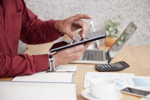 Pessoa utilizando um tablet enquanto trabalha em um ambiente de escritório, com notebook, calculadora, gráficos financeiros, bloco de notas e xícara de café sobre a mesa. A cena representa a conectividade e a análise de dados em tempo real, aspectos essenciais do conceito de Open Finance, que promove a integração segura de informações financeiras entre instituições, ampliando a oferta de serviços personalizados e inovadores no setor bancário.