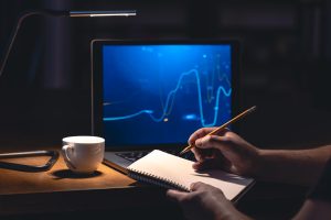 Pessoa segurando um caderno e um lápis, fazendo anotações enquanto observa gráficos financeiros exibidos na tela de um notebook, iluminado por uma luminária de mesa em um ambiente escuro. Ao lado, há uma xícara branca sobre a mesa, compondo um cenário de estudo e análise de fundos de investimento, com foco em interpretar tendências e flutuações do mercado para tomar decisões estratégicas e bem fundamentadas.
