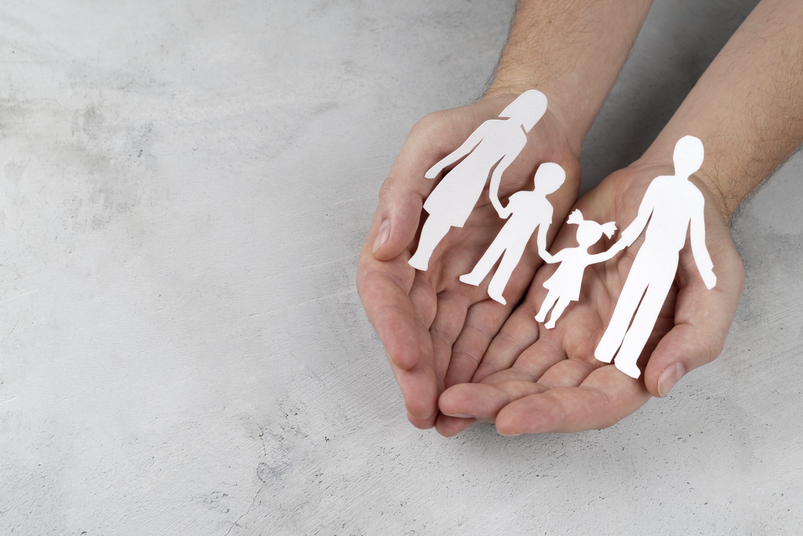 Mãos abertas segurando recortes de papel representando uma família composta por dois adultos e duas crianças, simbolizando proteção e cuidado. A imagem ilustra o conceito de segurança e amparo familiar, valores essenciais no mercado de seguros de vida, que oferece soluções para garantir estabilidade financeira e proteção aos entes queridos em situações imprevistas.