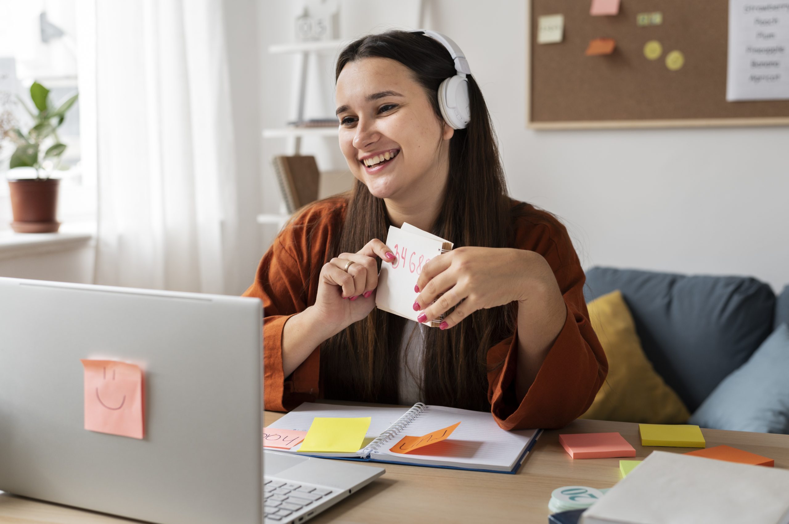 Mulher sorridente, usando fones de ouvido brancos, participa de uma videochamada enquanto segura cartões de anotações, sentada em um ambiente acolhedor de home office. À sua frente, há um notebook com post-its coloridos, um caderno aberto e materiais organizados sobre a mesa. Ao fundo, prateleiras com livros e plantas complementam o espaço, refletindo um estilo de trabalho remoto cada vez mais valorizado no mercado imobiliário, que busca adaptar os imóveis às novas necessidades de home office e produtividade em casa.
