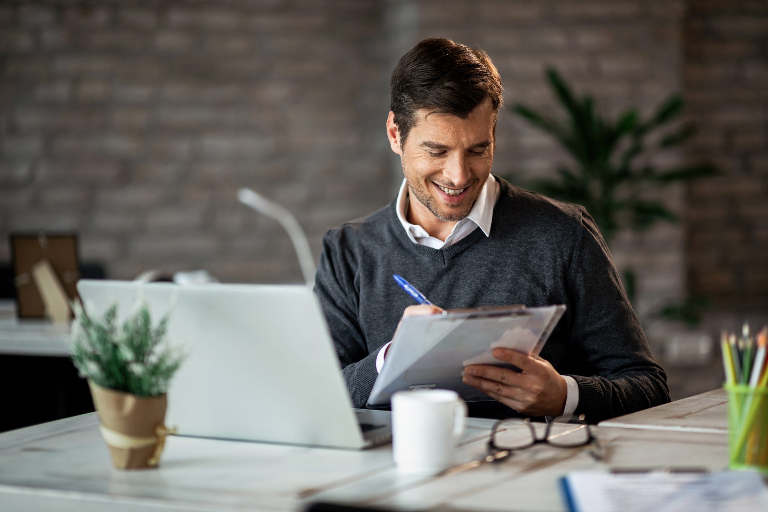 Homem sorridente, vestido com camisa branca e suéter cinza, sentado em uma mesa de escritório moderna enquanto revisa anotações em uma prancheta. Ao seu lado, há um laptop aberto, uma caneca branca e óculos de grau sobre a mesa. No ambiente ao fundo, decorado com plantas e iluminação suave, destaca-se um cenário profissional propício ao planejamento financeiro eficiente e estratégico.