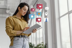 Mulher jovem com óculos e camisa mostarda utilizando um tablet em um ambiente iluminado, enquanto ícones gráficos de redes sociais como curtidas, notificações, mensagens e carrinho de compras flutuam ao seu redor, representando interações digitais e conectividade online.