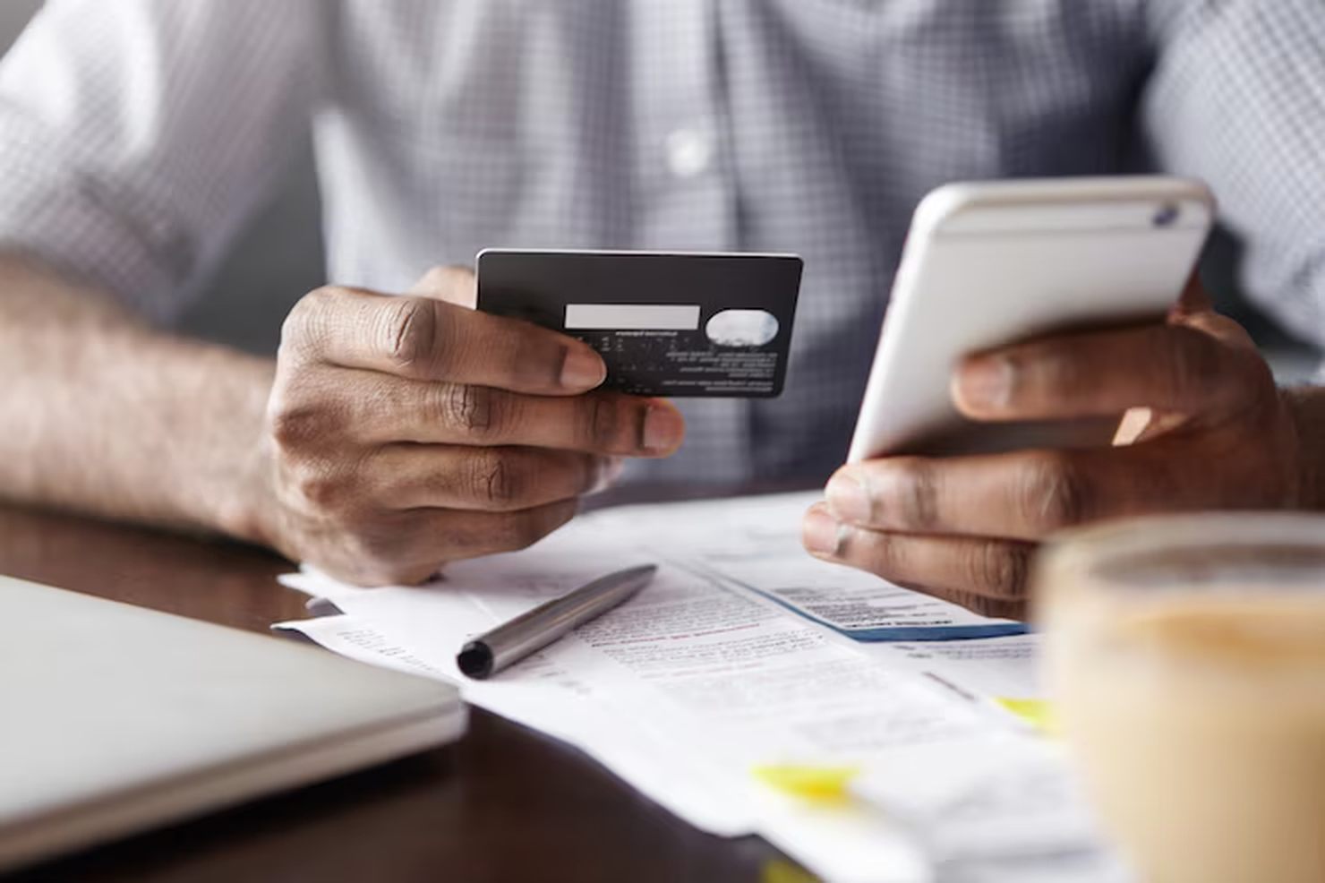 Pessoa segurando um cartão de crédito em uma mão e um smartphone na outra, diante de documentos e fatura sobre a mesa, representando a análise e gestão de finanças pessoais relacionadas ao crédito consignado.