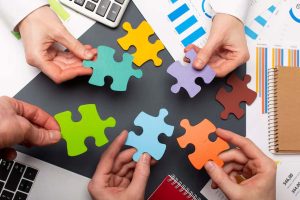 Diversas mãos segurando peças coloridas de quebra-cabeça sobre uma mesa com gráficos, cadernos e dispositivos eletrônicos, simbolizando integração de ideias e trabalho em equipe no contexto da economia colaborativa.