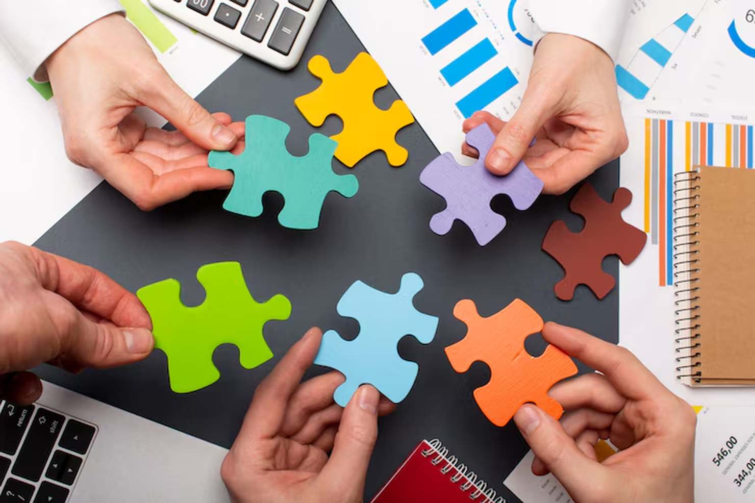 Diversas mãos segurando peças coloridas de quebra-cabeça sobre uma mesa com gráficos, cadernos e dispositivos eletrônicos, simbolizando integração de ideias e trabalho em equipe no contexto da economia colaborativa.