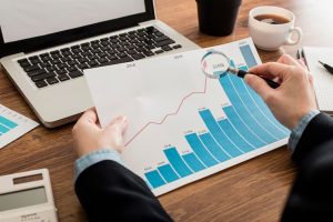 Pessoa analisa um gráfico de crescimento financeiro com uma lupa, evidenciando o aumento de rendimentos ao longo dos anos, com destaque para aplicações atreladas ao CDI, em um ambiente de escritório com notebook, calculadora e café sobre a mesa.