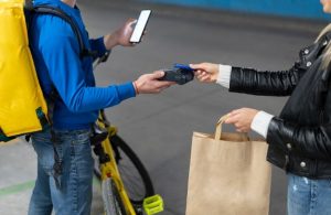 Entregador de aplicativo recebe pagamento por aproximação de uma cliente utilizando cartões de crédito com cashback em apps de delivery, durante a entrega de pedidos em sacola de papel reciclável.