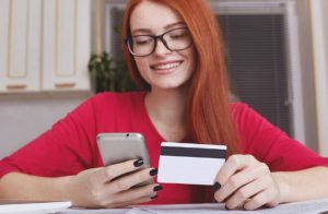 Mulher sorridente segura um celular e um cartão de crédito enquanto realiza uma transação online, refletindo o uso consciente e o controle sobre a anuidade do seu cartão de crédito no ambiente doméstico.