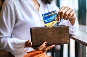 Mulher segura uma carteira aberta enquanto escolhe entre diferentes cartões de crédito nas cores azul, dourado e preto, representando as categorias cartões platinum, gold e black, com destaque para suas opções e benefícios exclusivos.