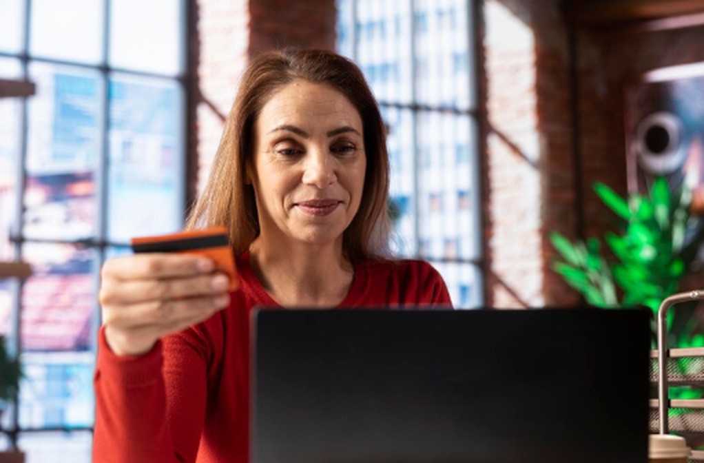 Mulher realiza uma compra online utilizando o notebook e segurando um cartão de crédito, simbolizando o uso do limite emergencial de cartão de crédito para concluir a transação em um ambiente doméstico iluminado.