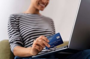 Mulher sorridente faz uma compra online usando notebook e cartão, representando o uso dos melhores cartões de crédito para viagens, que oferecem benefícios como milhas, pontos e isenção de taxas.