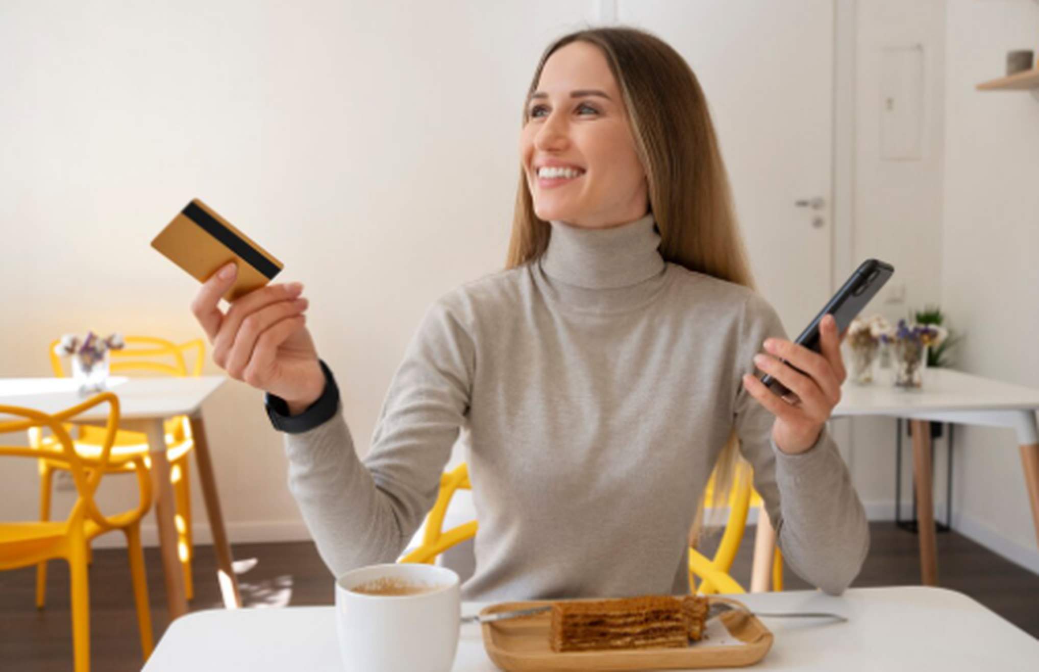 Mulher sorridente em uma cafeteria, segurando cartão e celular enquanto toma café, simbolizando praticidade no pagamento e as vantagens oferecidas por cartões de crédito com benefícios.