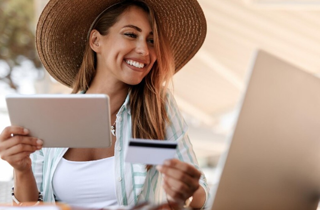 Mulher sorridente usando chapéu, segurando cartão de crédito e tablet enquanto faz compras online, ilustrando as vantagens de um programa de pontos flexível que permite acumular e trocar pontos em diferentes parceiros.