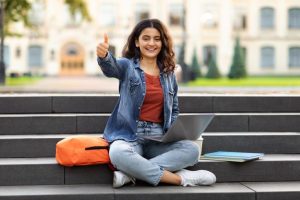 Estudante sorridente sentada em escadaria com mochila e laptop, mostrando sinal de positivo, representando os benefícios do Tesouro Educa+ no financiamento de estudos.