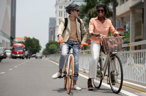 Duas pessoas andam de bicicleta em uma avenida movimentada, sorrindo e usando capacetes de segurança, representando um estilo de vida sustentável e o incentivo à mobilidade urbana.